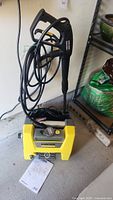 Front top view of the Karcher pressure washer showing the spray gun with attached hose standing upright, yellow and gray body, instruction manual on floor.