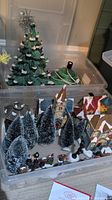 Ceramic green Christmas tree with white accents and silver star top, placed on plastic tote with cord and base, alongside resin village houses and winter trees inside a clear storage container.
