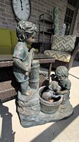 Full view of the resin statue showing a standing boy at a water pump and a kneeling girl with a dog inside a barrel, positioned on a concrete floor.