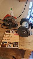 Side view of Black & Decker circular saw, Porter Cable bench grinder, and Woodworker's Manual on wooden surface.