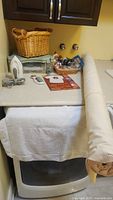 Wide view of partial bolt of creamy white upholstery fabric, Rowenta steam iron, baskets with sewing supplies on counter