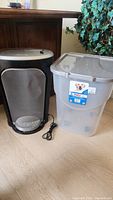Paper shredder and plastic storage bin side by side on floor against wooden furniture and plants.