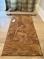 Full view of brown and gold tapestry with a lady sitting among large flowers, mounted on a gold resin hanging bar with finials.