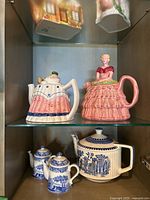 Five teapots displayed on glass shelving, featuring the HJ Wood Princess Charlotte teapot in pink, Cardinal Inc. picnic table teapot, and Spode and Sadler blue and white teapots