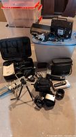 Lot of vintage cameras and accessories including Sony cameras, projection equipment, camera bags, and cords displayed on floor and storage bins
