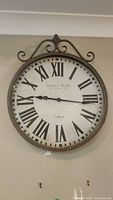 Front view of large round wall clock with Roman numerals and distressed metal frame with decorative top accent