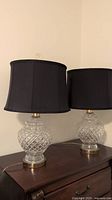 Full frontal view of both lamps showing crystal bases, brass-tone hardware and black drum shades, with one finial missing.