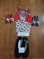 Top-down image showing the child's red silk dragon hat flanked by two pairs of children's dragon head shoes (red and blue pairs) and a pair of adult black slippers below, all arranged on wooden floor.