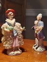 Two porcelain figurines, woman with basket of flowers and man with repaired bouquet arm, shown on wooden surface.
