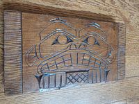 Wood carved panel depicting a stylized beaver face in First Nations design, showing natural wood texture and carved geometric pattern.