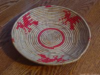 Angled side view of a round, shallow hand woven basket with natural and red fibers showing geometric patterns, placed on wooden floor.