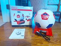 Front view of the Koolatron Coca Cola Soccer Ball Mini Fridge Cooler with power cord and manual beside it, box in background.
