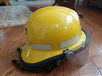 Side view of yellow vintage Cairns & Bros firefighter helmet with face shield and chin strap on wooden floor.