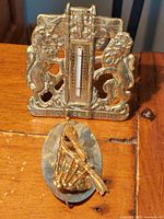 Both the brass coat of arms plaque and the gold plated bagpipe kilt pin positioned on a wooden surface. Coat of arms shows two heraldic lions and a built-in thermometer which is broken.