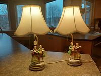 Photo showing both porcelain figural lamps with textile shades and brass bases placed on countertop with window in background.