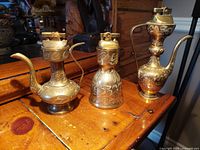 Three brass and metal vintage lighters placed on a wooden surface, including two genie lamp styles and a rounded form lighter.