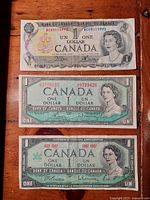 Three Canadian one dollar bills laid out on a wooden surface showing front designs including Queen Elizabeth II portraits and serial numbers for two of the bills.