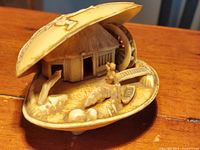 Photo showing the clam shell diorama open, revealing the carved interior with house, water wheel, figure, and animals