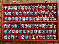 Front view of ninety miniature hand painted Eastern Asian theatre masks mounted neatly on a red fabric covered display board with gold trim and hanging cord.