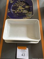 Photo showing one dark blue square plate with gold and floral motif and white rectangular ceramic trays beneath