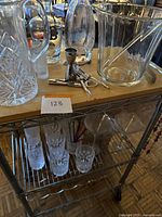 Overall view of metal and wood top bar cart with glassware and bar tools on top shelf and glasses on middle shelf