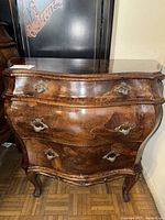 Front view of burlwood chest showing three drawers with ornate metal handles and curved legs.
