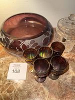 Five small grape carnival glasses with iridescent finish, purple etched rounded bowl with floral patterns and chip on rim, clear faceted crystal lidded jar.