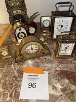 Photo showing eight small clocks in various styles including brass, metal, and decorative painted wood on marble surface.