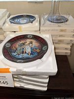 Stack of decorative plates in protective white foam cases on table, showing colorful classical scene on top plate.
