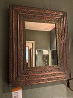 Front view of copper toned rectangular mirror with ornate patterned frame hung on dark textured wall.
