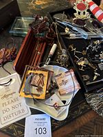 Wide shot showing most items spread out on black table surface. Visible items include pens in wood box, cuff links arranged on tray, eyeglasses, Algonquin Hotel sign, picture frame, stamped silver choker, and other small charms.