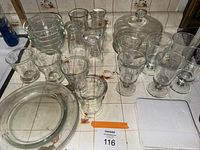 Photo of multiple glass items including tumblers, footed glasses, bowls, plates, and lidded cake plate on tiled surface.
