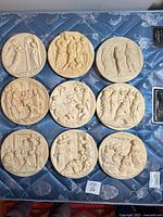 Top view of all nine circular carved plates laid out on blue quilted fabric showing detailed classical and biblical scenes