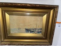 View of full framed artwork showing seascape with boats and dock, behind glass, in gold-tone ornate frame.