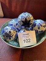 Set of five blue and white painted porcelain orbs arranged inside a large floral patterned porcelain bowl, shown on a couch.