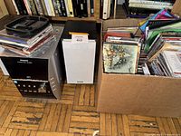 Wide shot showing Panasonic stereo system, a pair of speakers, and a box full of CD cases with some CDs stacked on top of the stereo.
