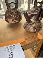 Three Peruvian double spout vessels with detailed painted decorations featured on a wooden surface with window background