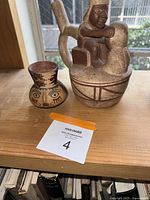 Two Peruvian pottery items shown together on a wooden shelf, including a larger figure with a man and a smaller vessel with a human face design.