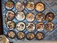 Seventeen 8-inch collectible plates laid out on a blue patterned cloth, showing various detailed scenes of Russian life, landscapes, and architecture.