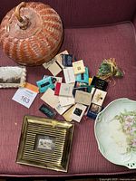 Photo of woven basket with lid, glass paperweight, floral plate, and assorted matchbooks on a red upholstery surface.