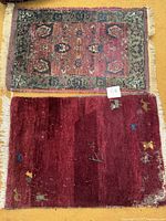 Photo showing the two small rectangular rugs stacked, one deep red with small motifs and one with floral geometric design with a green border