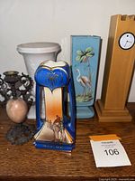 Photo showing a group of decorative items including the Noritake blue vase with camel scene, white milk glass urn, light blue flamingo urn, wooden tower clock, metal candle holder and beige vase.