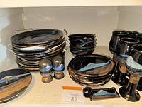 Stacked ceramic dinnerware including 10-inch plates, bowls, side plates, goblets, salt and pepper shakers, and a spoon rest all with black glaze and blue, brown, and white brush strokes.