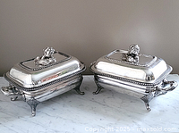 Front view of two silver-plated covered entrée dishes on warming trays atop marble surface.