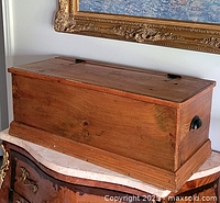 Front view of the large antique wooden box sitting atop a piece of furniture under a framed painting. Shows rectangular box with hinged lid and natural wood grain.