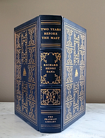 Front and spine view showing dark blue genuine leather cover with gold embossed intricate floral and scroll designs, including title and author on spine, featuring raised spine hubs.