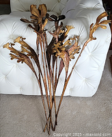 Photo showing a group of large artificial plant stems with brown and gold leaves, arranged on a white tufted surface.