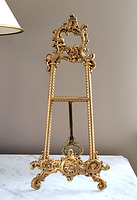 Front full view of the gold ornate metal easel stand showing its detailed baroque-style decoration and sturdy curved feet.
