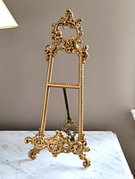 Front view of gold ornate metal easel stand on marble surface showing decorative design and measuring approximately 15.5 inches in height.