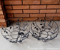 Two round metal hanging basket frames with leaf pattern and chains, resting on concrete near brick wall.